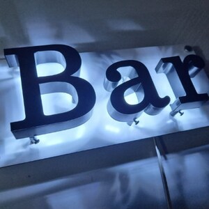 May include: A white sign with the word "Bar" in black, raised, 3D letters. The letters are illuminated from behind, creating a bright white glow.