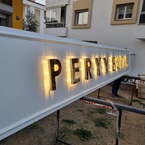 May include: A white sign with the words "PERY BUTIK" in gold letters with lights behind them.