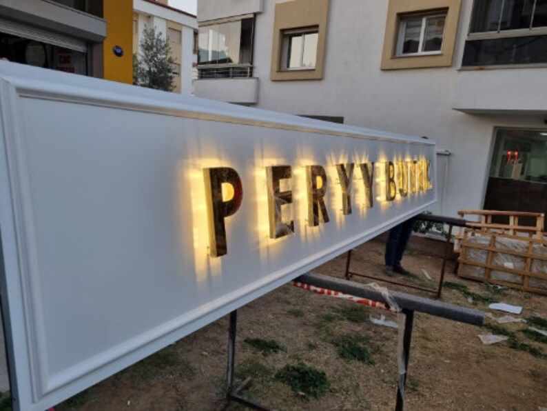May include: A white sign with the words "PERY BUTIK" in gold letters with lights behind them.