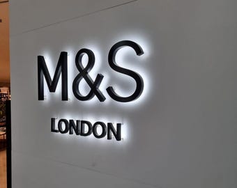 Puede incluir: Un letrero de pared con las letras negras M&S y la palabra LONDON situada justo debajo en un tamaño de fuente menor. Las letras están retroiluminadas con un brillo blanco intenso, creando un efecto de halo sobre la pared blanca.