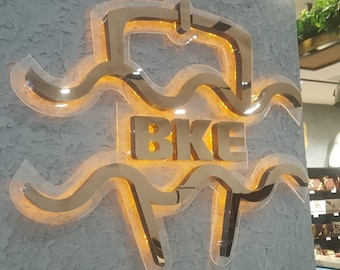 May include: A gold-coloured sign with the word "BKE" in large letters, surrounded by a wave-like design. The sign is illuminated from behind, creating a warm glow.