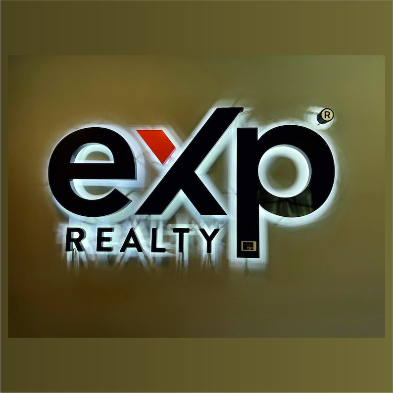 May include: A white sign with black lettering that reads "exp REALTY" with a red accent in the "x" and a registered trademark symbol in the top right corner.