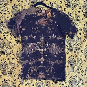 Pode incluir: Uma camiseta escura tie-dye com um padrão simétrico único em tons de preto, marrom e roxo. A camisa está pendurada em um cabide preto em um fundo amarelo estampado. A camiseta de manga curta tem decote em V.