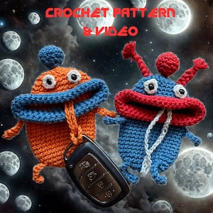 May include: Two crocheted alien-like figures, one orange and blue, the other blue and red, hold hands. A car key fob hangs from the orange figure. The background features moons and the text "CROCHET PATTERN & VIDEO".