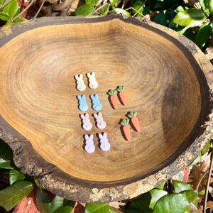 May include: A wooden bowl filled with pastel colored bunny and carrot shaped earrings. The earrings are made of polymer clay and have a glossy finish.