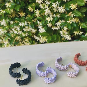 May include: Five pairs of floral hoop earrings in black, lavender, and coral. The earrings are displayed on a white surface.