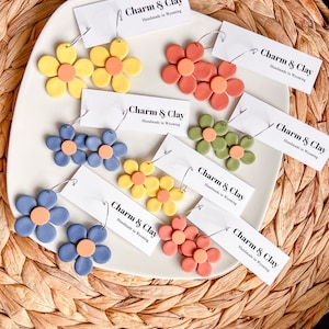 May include: Assortment of handmade polymer clay earrings in various colors, including yellow, orange, red, green, blue, and brown. Each earring features a flower design with a small circle in the center. The earrings are displayed on a white surface with a tag that reads "Charm & Clay Handmade in Wyoming".
