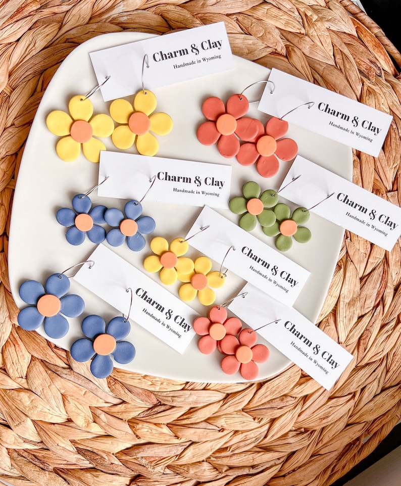 May include: Assortment of handmade polymer clay earrings in various colors, including yellow, orange, red, green, blue, and brown. Each earring features a flower design with a small circle in the center. The earrings are displayed on a white surface with a tag that reads "Charm & Clay Handmade in Wyoming".