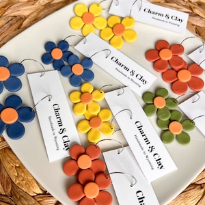 May include: Assortment of colorful clay flower earrings with hoop backs. The earrings are displayed on a white plate with a tag that reads "Charm & Clay Handmade in Wyoming".
