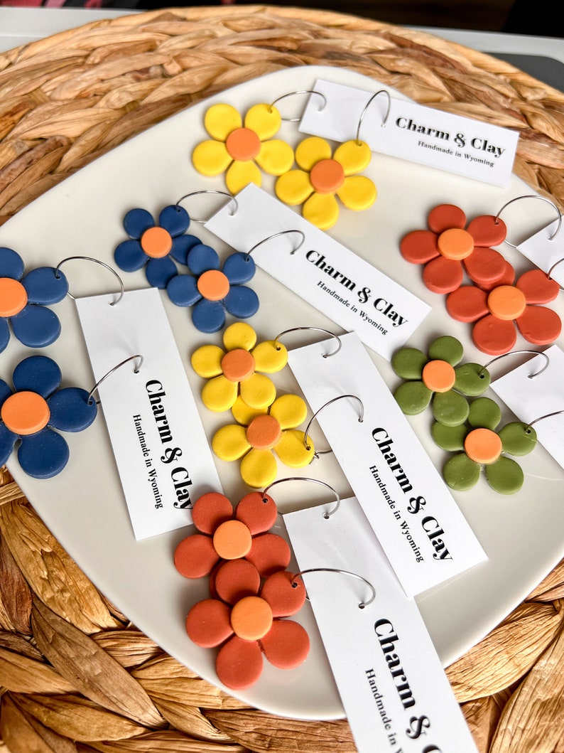 May include: Assortment of colorful clay flower earrings with hoop backs. The earrings are displayed on a white plate with a tag that reads "Charm & Clay Handmade in Wyoming".