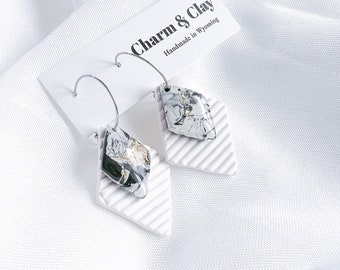 May include: White ceramic diamond-shaped earrings with a black and white marbled design and gold accents. The earrings are on a white card with the text "Charm & Clay Handmade in Wyoming".