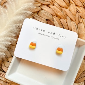 May include: A pair of candy corn earrings made of polymer clay. The earrings are yellow, orange, and white. The earrings are on a white card with the text "Charm and Clay Handmade in Wyoming".