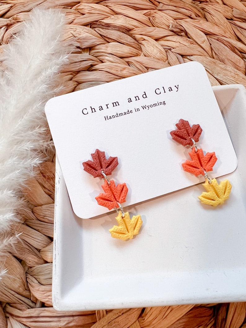 May include: A pair of dangle earrings featuring three layered maple leaves in shades of red, orange, and yellow. The earrings are handmade in Wyoming and are labeled "Charm and Clay".