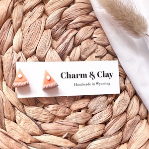 Può includere: Un paio di orecchini a forma di fetta di torta di zucca, di colore marrone e bianco, su una carta bianca con il testo "Charm & Clay Handmade in Wyoming".
