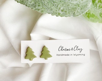 May include: A pair of green clay earrings shaped like Christmas trees. The earrings are displayed on a white card with the text "Charm & Clay Handmade in Wyoming".