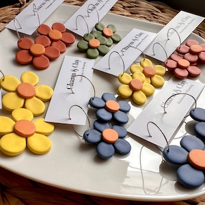 May include: Assortment of handmade polymer clay earrings in various colors, including yellow, orange, red, blue, and green. Each earring features a flower design with a center circle. The earrings are displayed on a white plate with a business card that reads "Charm & Clay Handmade in Wyoming".
