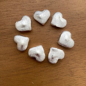 K&ouml;nnte beinhalten: Sieben silberne herzf&ouml;rmige Anh&auml;nger, die in einem Kreis auf einer braunen Holzoberfl&auml;che angeordnet sind.