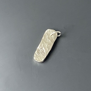 May include: A silver pendant with a hammered texture and an engraved symbol. The pendant is rectangular and has a loop at the top for attaching to a chain.