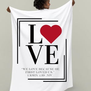 以下が含まれることがあります： 大きな黒い文字で「LOVE」という言葉と赤いハートが描かれた白いブランケット。ブランケットには、1ヨハネ4:19（NIV）からの引用「WE LOVE BECAUSE HE FIRST LOVED US.」も黒い文字で書かれています。