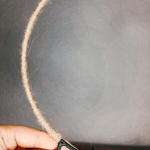 May include: A close-up of a hand holding a brown rope wrapped around a black metal hoop. The rope is being wrapped around the hoop to create a wreath.