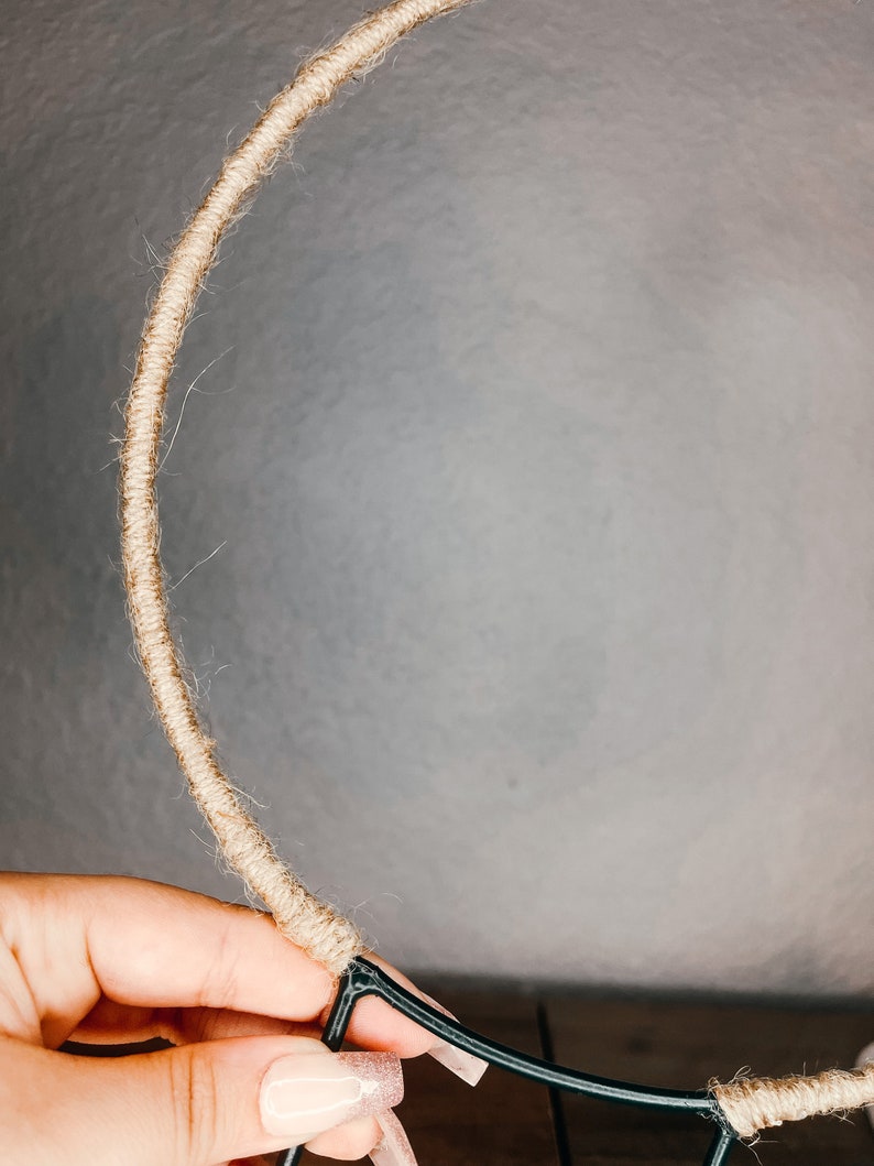 May include: A close-up of a hand holding a brown rope wrapped around a black metal hoop. The rope is being wrapped around the hoop to create a wreath.