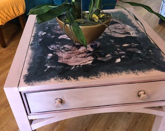 May include: A pink side table with a drawer and a gold planter with a green orchid plant on top. The table has a black and white patterned surface with the word "GERALD" visible.
