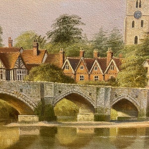 May include: A painting of a stone bridge with three arches over a river. The bridge is in front of a row of houses with red tile roofs. A tall church with a pointed spire is in the background.