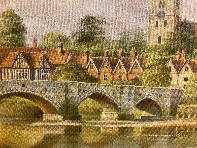 May include: A painting of a stone bridge with three arches over a river. The bridge is in front of a row of houses with red tile roofs. A tall church with a pointed spire is in the background.