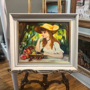 Puede incluir: Una pintura de una mujer con un sombrero amarillo y un vestido blanco, sentada en un jard&iacute;n con una cesta de fruta. La pintura est&aacute; enmarcada en un marco blanco.