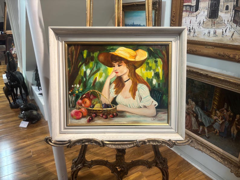 Puede incluir: Una pintura de una mujer con un sombrero amarillo y un vestido blanco, sentada en un jard&iacute;n con una cesta de fruta. La pintura est&aacute; enmarcada en un marco blanco.