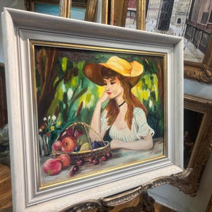 Puede incluir: Una pintura de una mujer con un sombrero amarillo y un vestido blanco, sentada en un jard&iacute;n con una cesta de fruta. La pintura est&aacute; enmarcada en un marco blanco.
