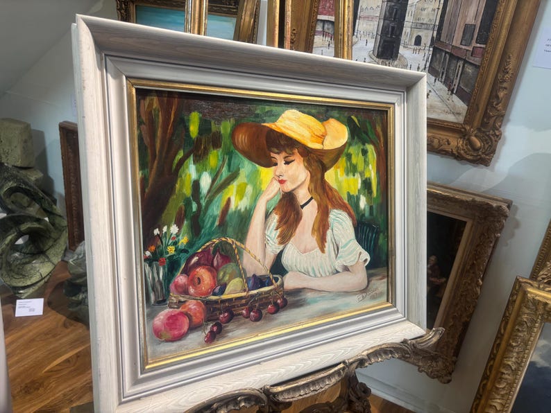 Puede incluir: Una pintura de una mujer con un sombrero amarillo y un vestido blanco, sentada en un jard&iacute;n con una cesta de fruta. La pintura est&aacute; enmarcada en un marco blanco.
