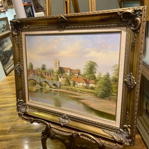 May include: A framed oil painting depicting a picturesque English village scene with a stone bridge over a river, a church in the background, and lush greenery.