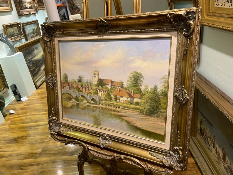 May include: A framed oil painting depicting a picturesque English village scene with a stone bridge over a river, a church in the background, and lush greenery.