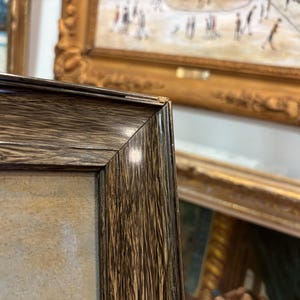 May include: Close-up of a dark brown wooden picture frame with a light brown canvas inside. The frame has a smooth, polished finish and a slightly raised edge.