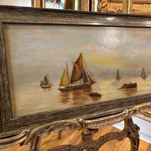 May include: A framed oil painting depicting a seascape with multiple sailboats on a calm body of water. The sky is a soft blue with hints of pink and yellow, and the water is a light blue with reflections of the boats. The painting is framed in a dark wood frame.
