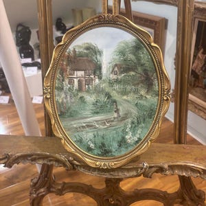 May include: A small oval painting in a gold frame depicts a cottage with a white picket fence and a path leading to it. The painting is set against a backdrop of green trees and a blue sky. There are white birds in the foreground.