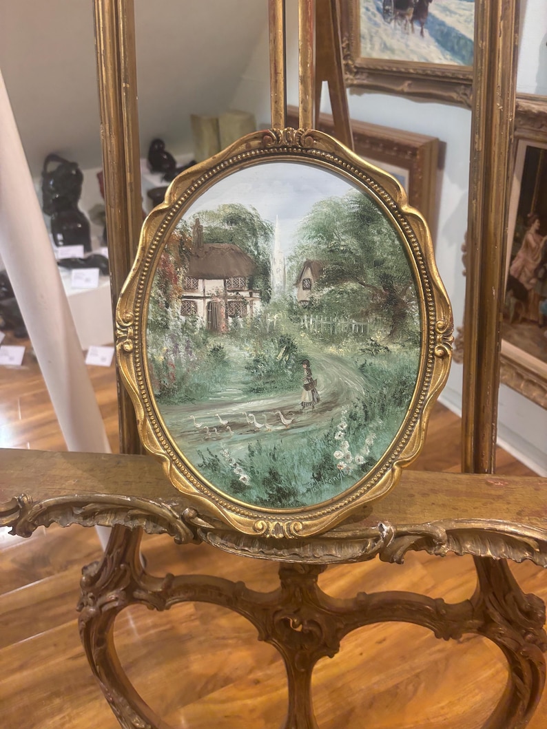 May include: A small oval painting in a gold frame depicts a cottage with a white picket fence and a path leading to it. The painting is set against a backdrop of green trees and a blue sky. There are white birds in the foreground.
