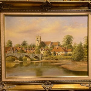 May include: A framed oil painting depicting a picturesque English village scene. The painting features a stone bridge over a river, with quaint houses and a church tower in the background. The sky is a soft, pastel blue with hints of pink and orange.