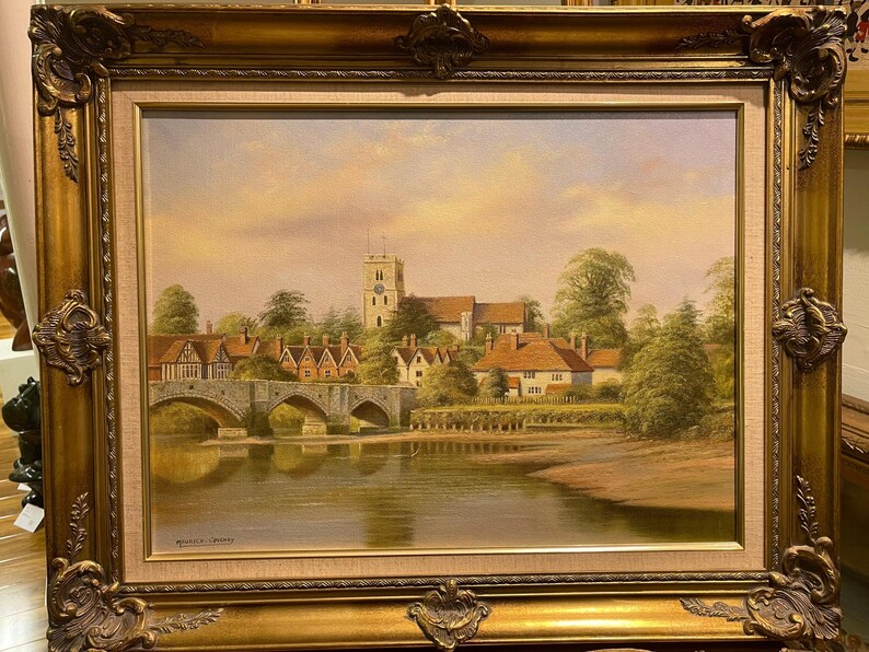 May include: A framed oil painting depicting a picturesque English village scene. The painting features a stone bridge over a river, with quaint houses and a church tower in the background. The sky is a soft, pastel blue with hints of pink and orange.