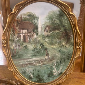 May include: An oval painting framed in gold, depicting a thatched cottage nestled amongst trees and a winding path. A woman in a black hat and dress walks along the path with a basket. The painting has a light blue sky with white clouds.