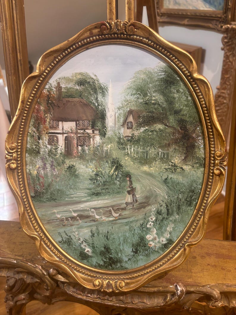 May include: An oval painting framed in gold, depicting a thatched cottage nestled amongst trees and a winding path. A woman in a black hat and dress walks along the path with a basket. The painting has a light blue sky with white clouds.