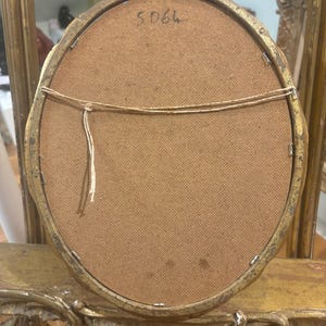 May include: The back of a gold oval picture frame with a brown fabric backing. The number "5066" is written on the back of the frame.