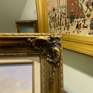 May include: A close-up of a gold-colored ornate picture frame with a detailed design. The frame is partially visible and is next to another picture frame.