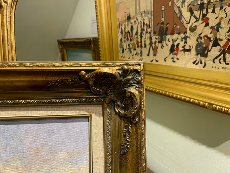 May include: A close-up of a gold-colored ornate picture frame with a detailed design. The frame is partially visible and is next to another picture frame.