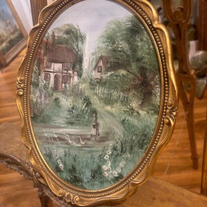 May include: A framed oval painting depicting a cottage in a verdant landscape, with a woman walking a flock of white geese. The painting is set in a gold frame with an ornate border.