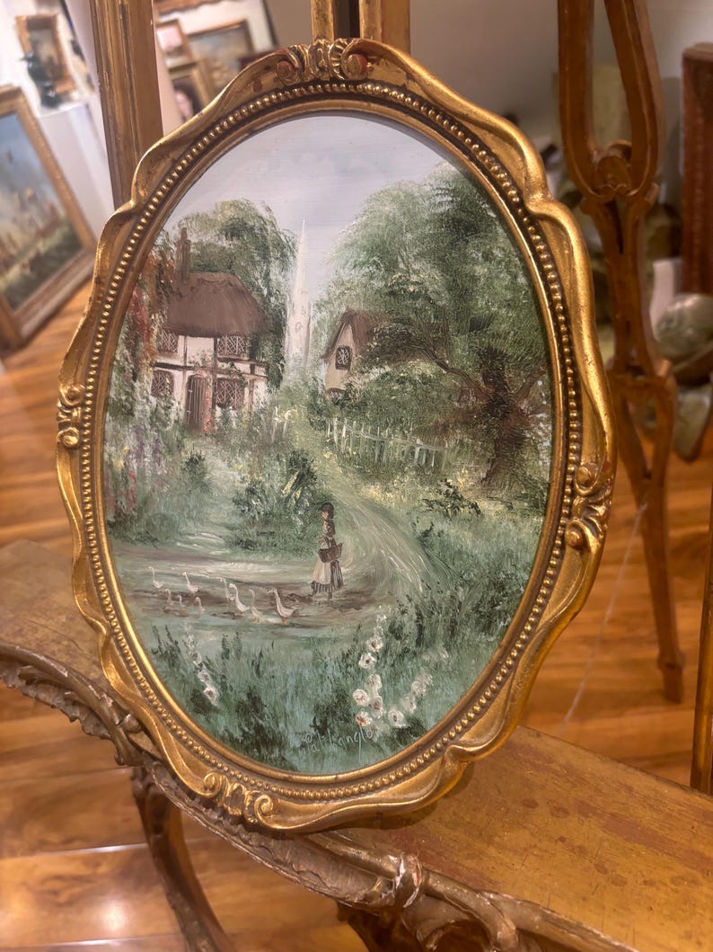May include: A framed oval painting depicting a cottage in a verdant landscape, with a woman walking a flock of white geese. The painting is set in a gold frame with an ornate border.