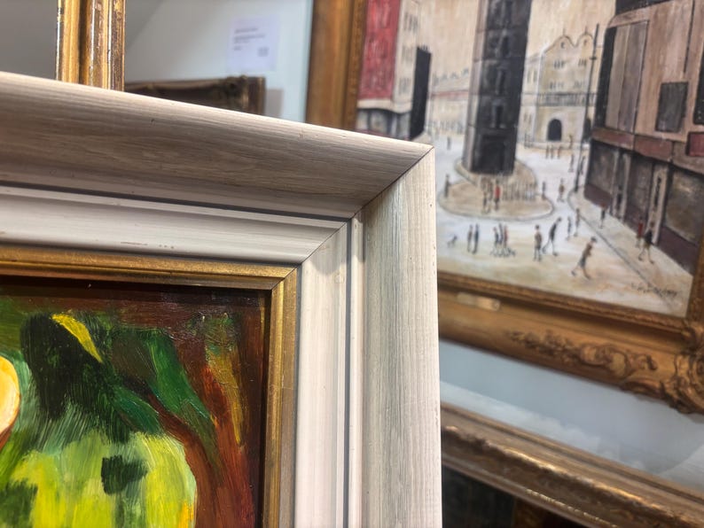 Puede incluir: Una pintura enmarcada con un marco blanco y dorado. La pintura representa una naturaleza muerta con una fruta verde y amarilla y un fondo marr&oacute;n.