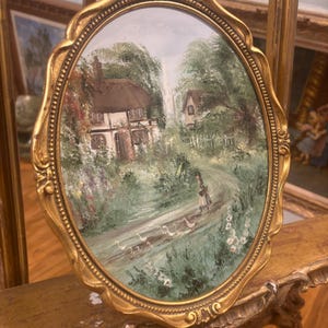 May include: An oval painting framed in gold, depicting a thatched cottage surrounded by trees and a path with a woman and geese. The frame has ornate details.