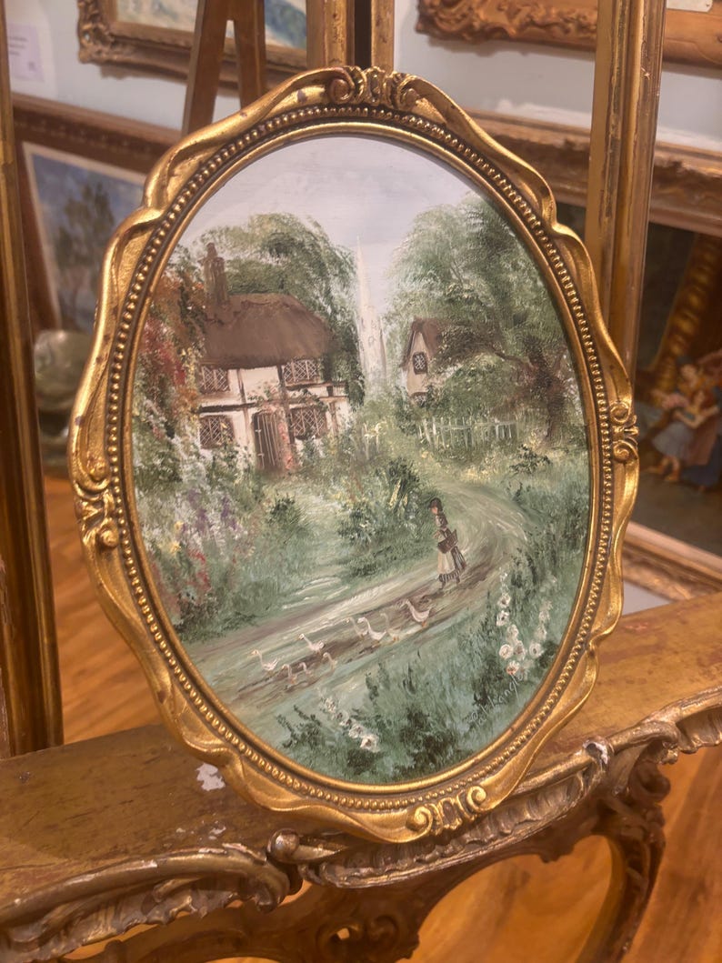 May include: An oval painting framed in gold, depicting a thatched cottage surrounded by trees and a path with a woman and geese. The frame has ornate details.